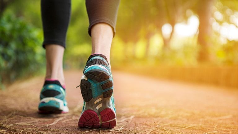 Close-up of a person walking or jogging on a path at sunrise, with sunlight shining through grass along the roadside.
