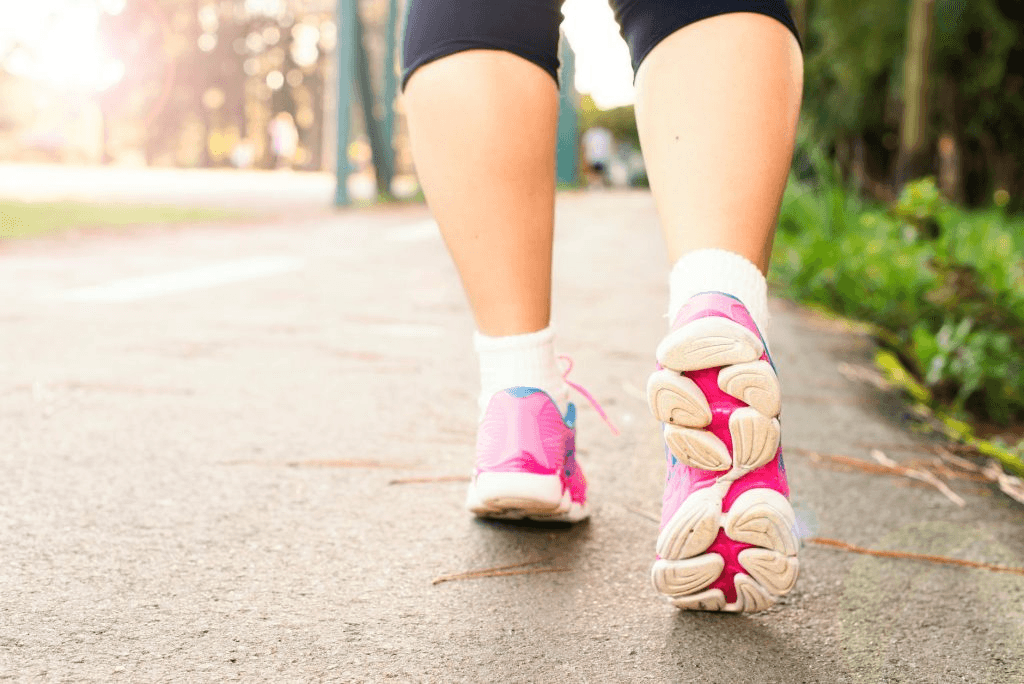 Close-up of a person walking or jogging on a path at sunrise, with sunlight shining through grass along the roadside.