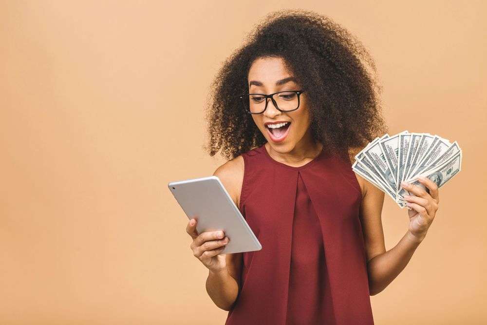 Image showing a happy woman using a tablet and holding a stack of dollar bills.