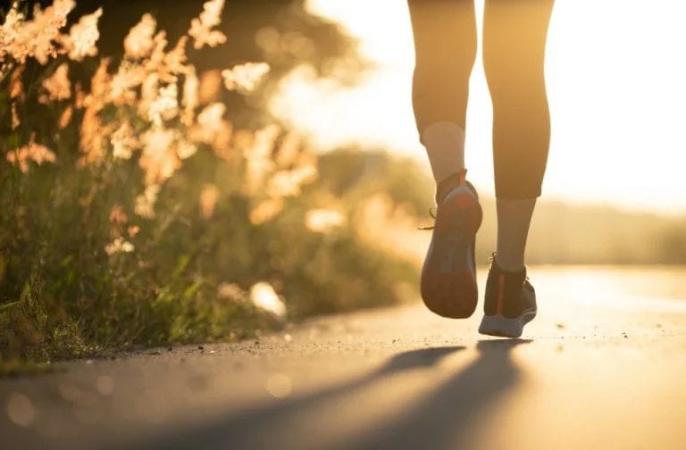 Close-up of a person walking or jogging on a path at sunrise, with sunlight shining through grass along the roadside.